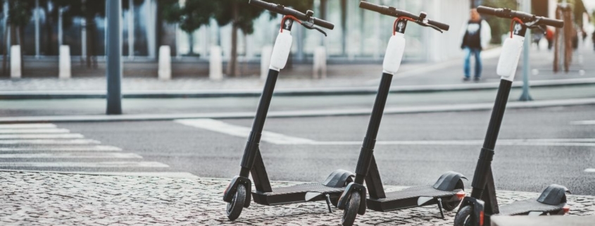 Photo of three electric scooters parked on a curb next to an empty intersection.