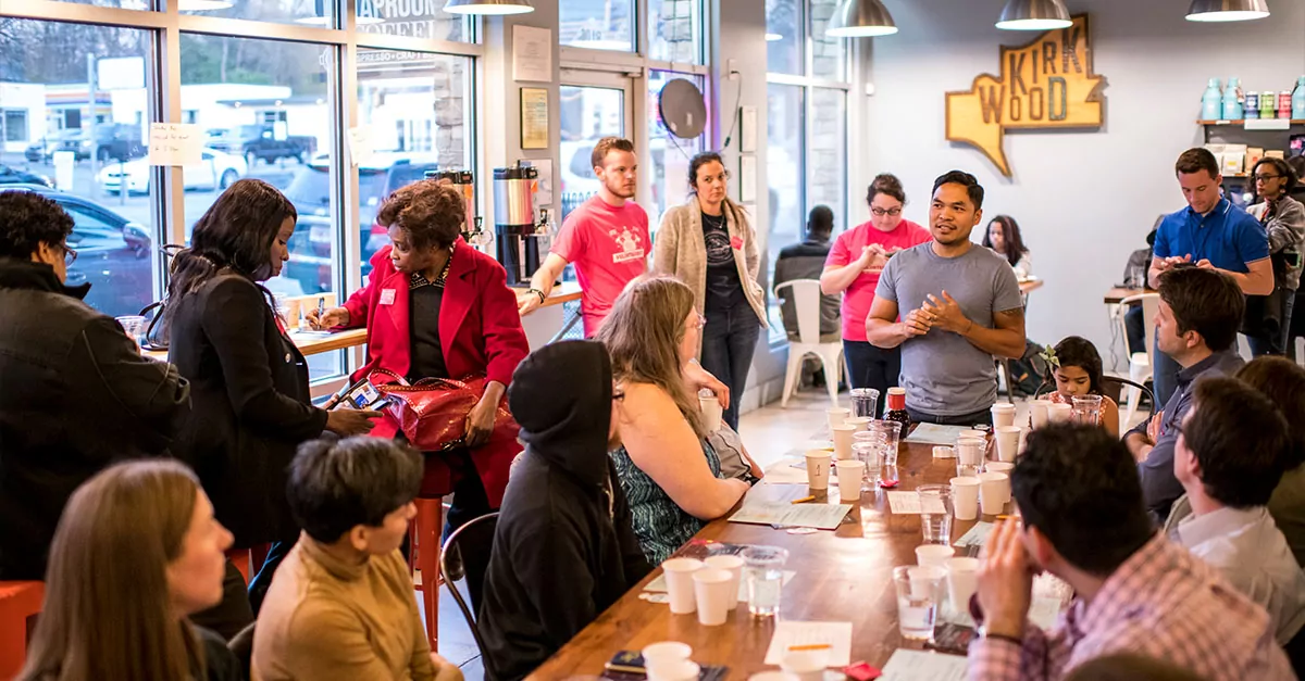 Lots of people gathered with coffee, listening to someone speak.